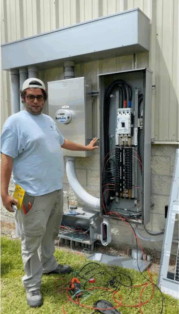 A man stands next to an open Vero Beach electrical panel and transfer switch setup outside a building, pointing towards the equipment. Electrical wires are visible at the base of the installation.