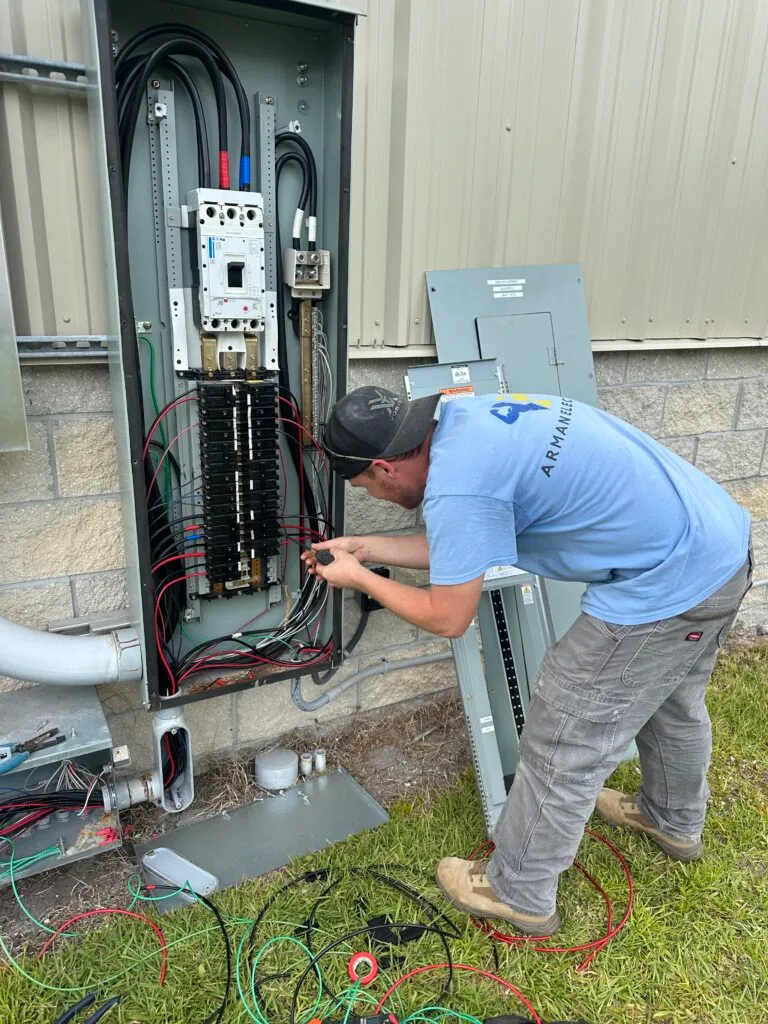 A male electrician wearing a blue t-shirt, a cap, and work pants, is working on an outdoor electrical panel or breaker box attached to the side of a building, with wires and tools scattered on the ground.